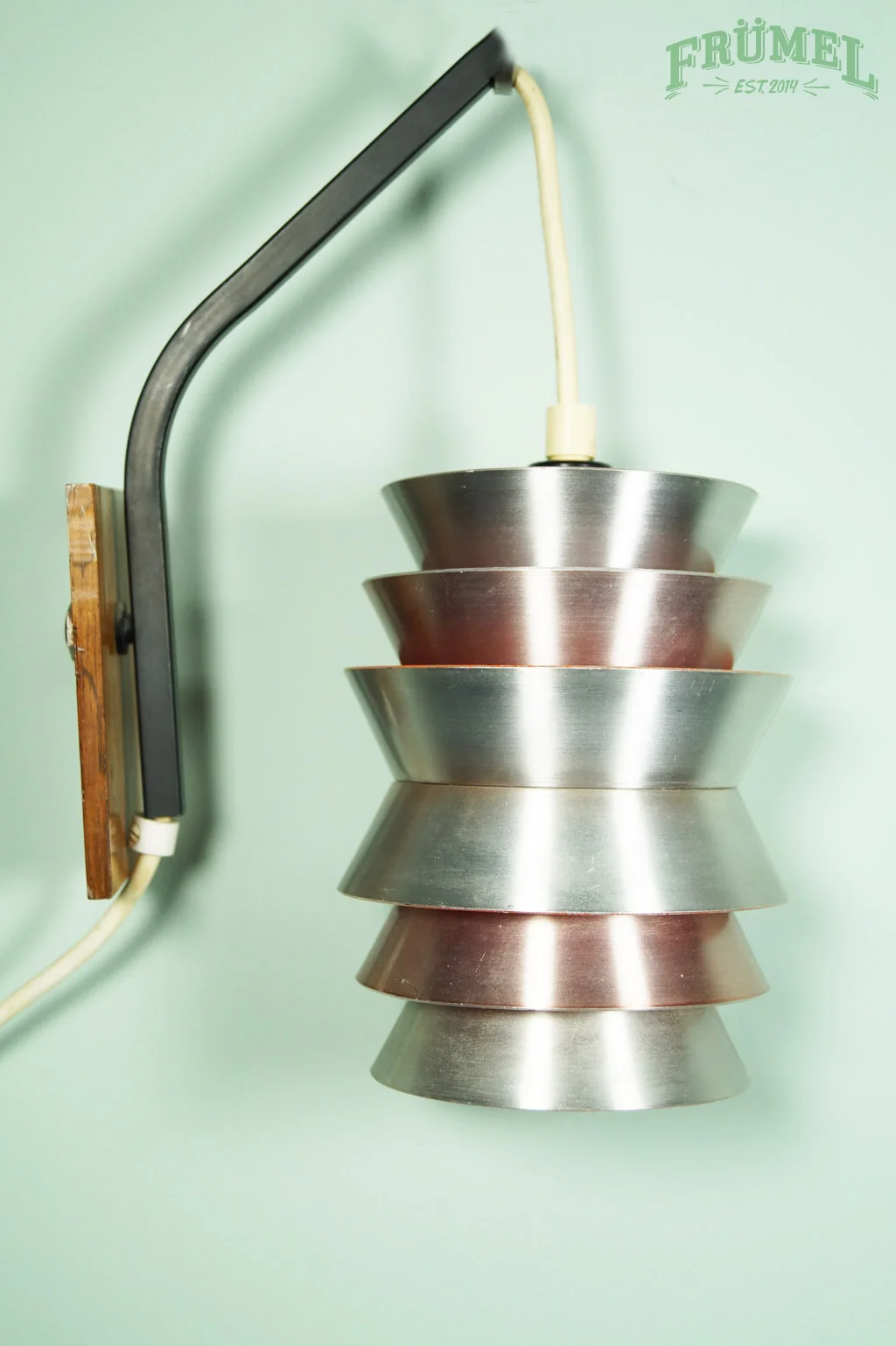 Decoratieve lamp met metalen schalen hangend aan een zwart metalen frame, bevestigd aan een groene muur met een logo "FRÜHEL EST.2014" in de boven rechterhoek.
