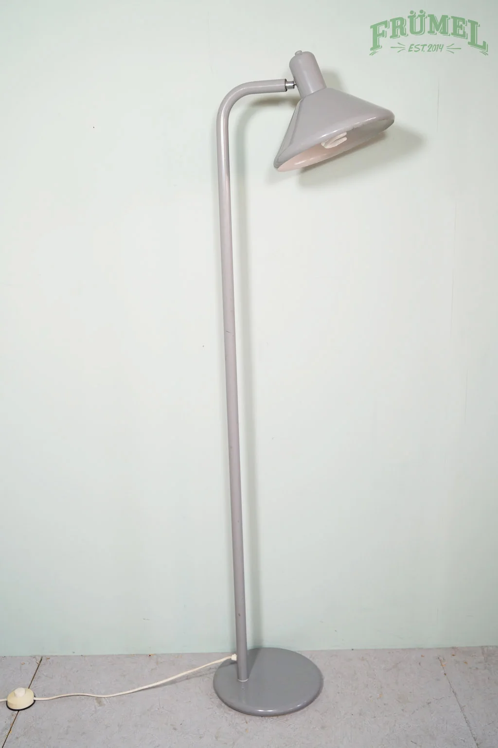 Een grijze staande lamp met verstelbare lampenkap, geplaatst op een lichte vloer tegen een lichtgroene muur met een groene opdruk van de tekst 'FRÜMEL EST.2014'.