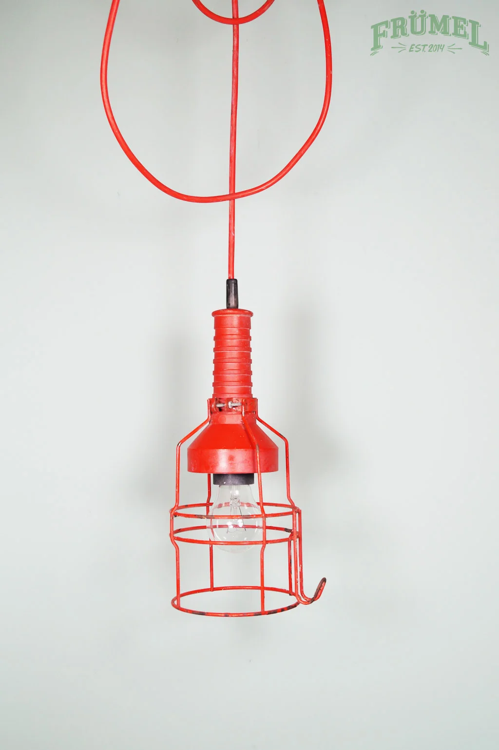 Een rode plafondlamp met een metalen veiligheidsrooster rondom de gloeilamp, hangend aan een draad, tegen een lichte muur.