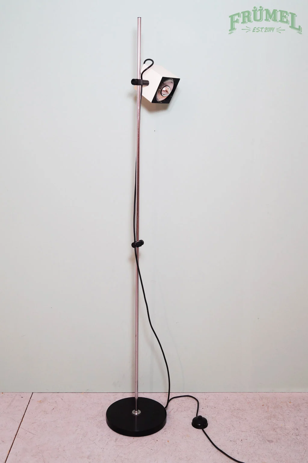 Staande lamp met een metalen pole, zwarte voet, en een hoekige lampenkap die naar beneden wijst, voor een witte achtergrond met groene tekst 'FRÜML' en 'EST. 2014' op de bovenhoek recht.