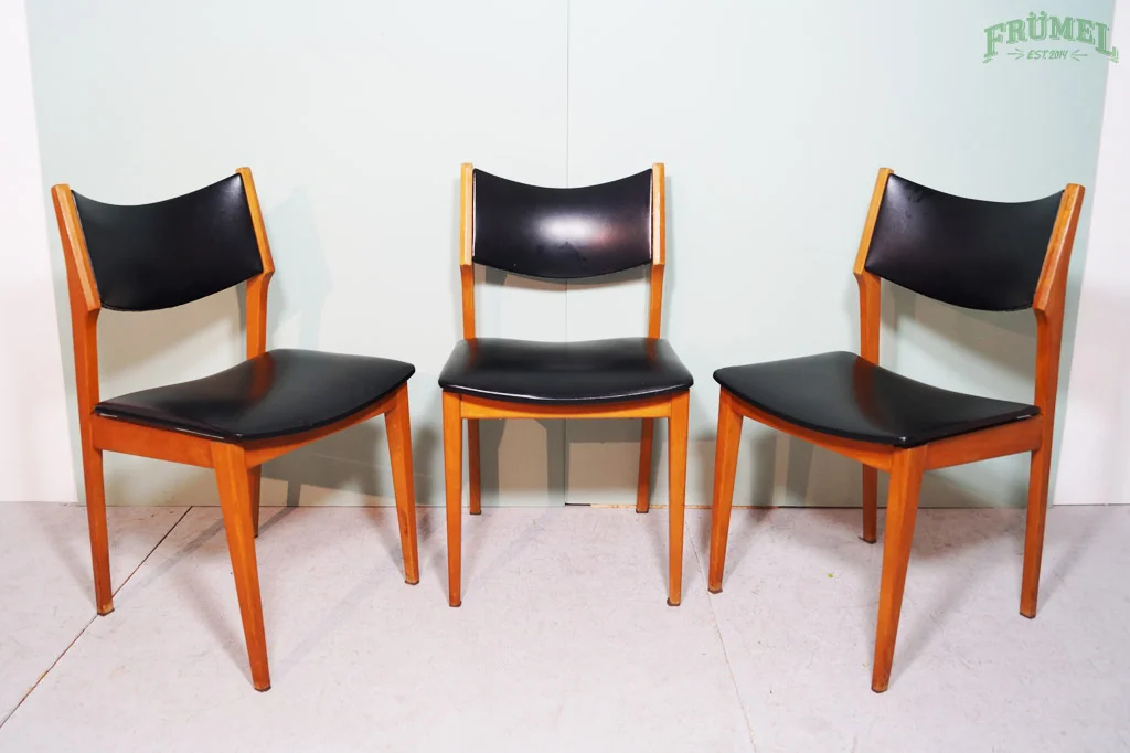 Drie houten stoelen met zwarte zittingen en rugleuningen, geplaatst tegen een witte muur.