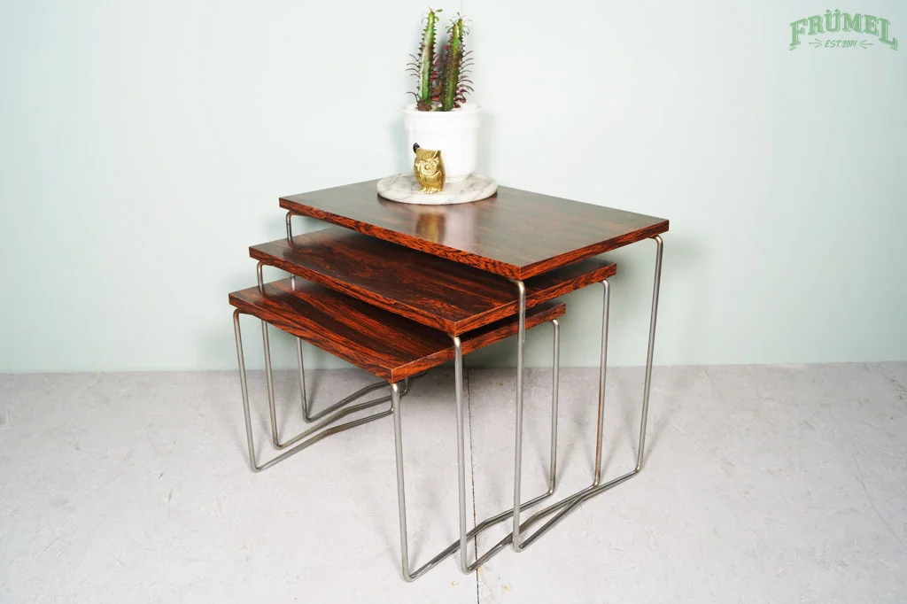 Set van drie houten bijzettafels met metalen poten, met een witte plantpot en een gouden uilbeeldje op de bovenste tafel, tegen een lichtgroene muur.