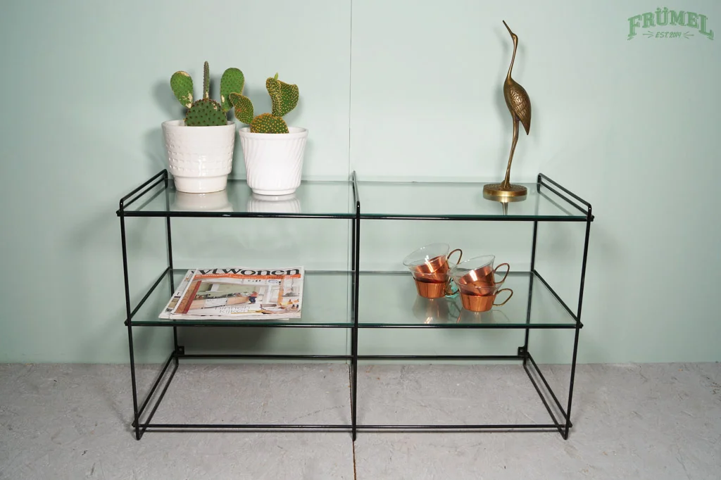 Wandplank met twee planten, een boek, koperen bekers, een gouden beeld en een krant in een lichte kamer.