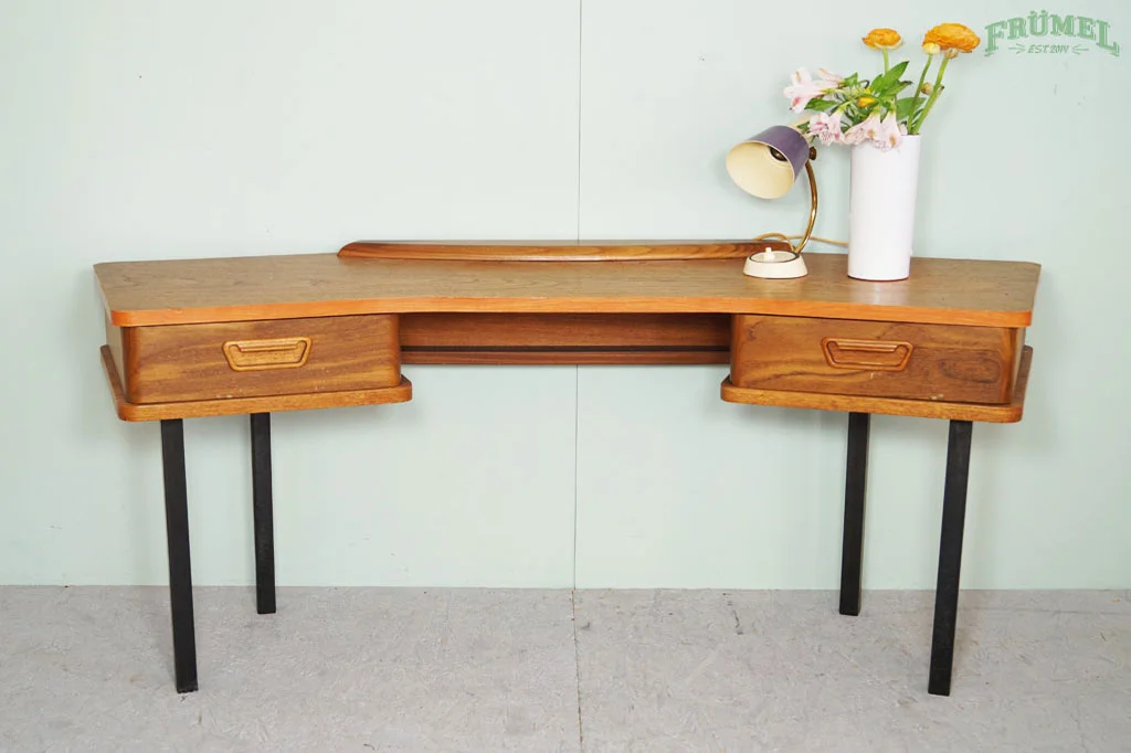 Een houten bureau met twee lades en zwarte poten, met een witte vaas met bloemen en een kleine bureaulamp erop, tegen een lichtgroene muur