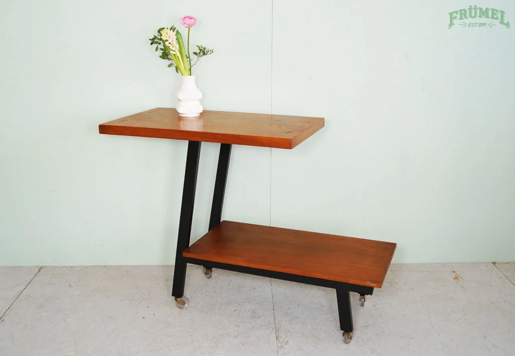 Een houten tafel met twee niveaus en een witte vaas met bloemen op de bovenste plank, tegen een lichtgroene muur met het tekstlogo 'FRÜMEL'.