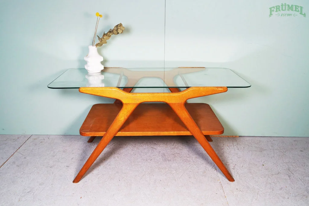 Vintage salontafel met een houten onderkant en een glazen bovenkant, met een witte vaas en gedroogde bloemen erop, tegen een lichtgroene muur.