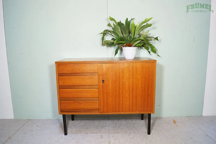 Houten dressoir met vier horizontale lades en een klepdeur, met een grote groene plant in een witte pot op top, tegen een lichtgroene muur.