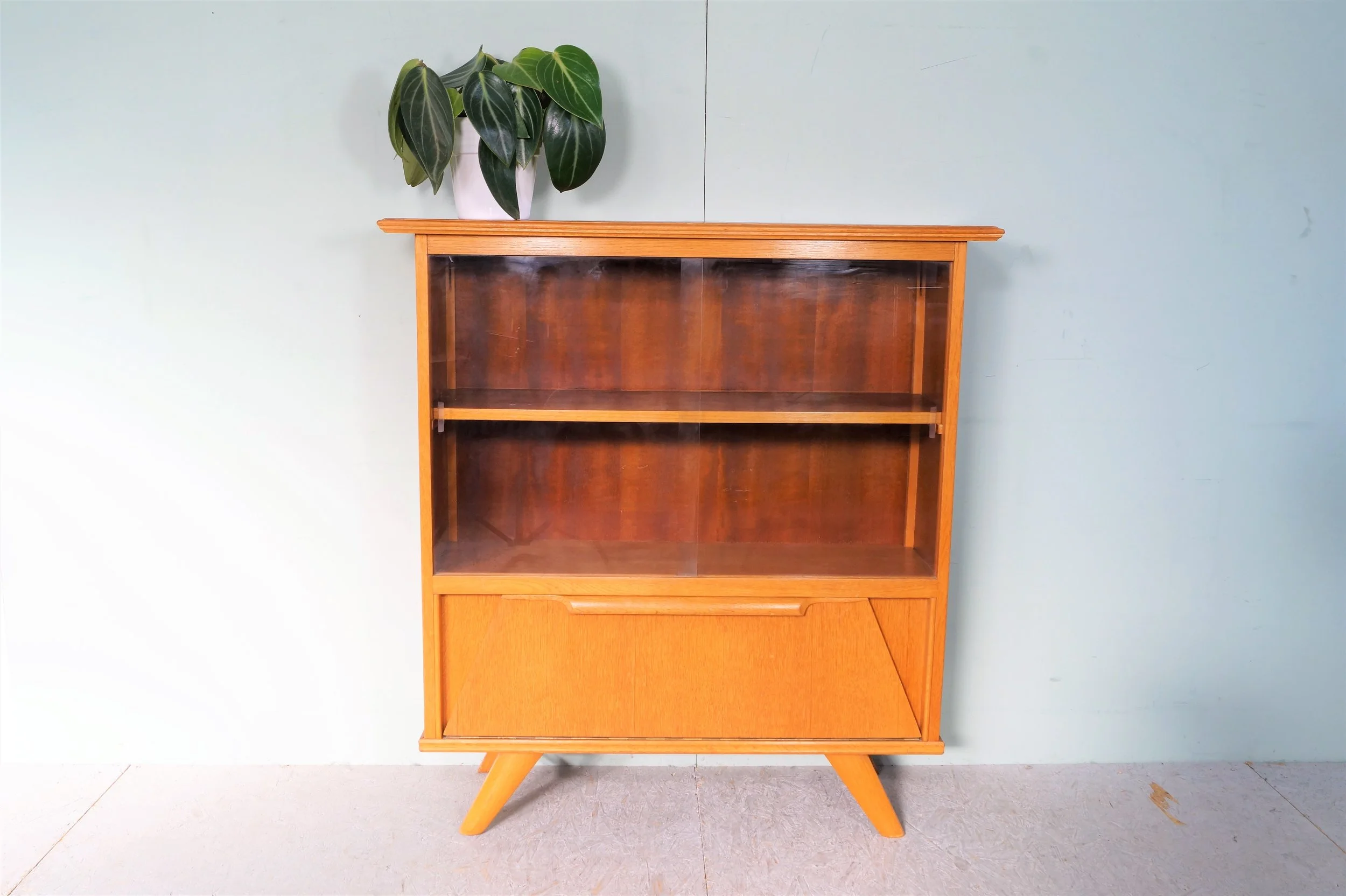 Vintage houten displaykast met glazen deur en een plant op top, geplaatst op een lichte vloer tegen een lichtblauwe muur.
