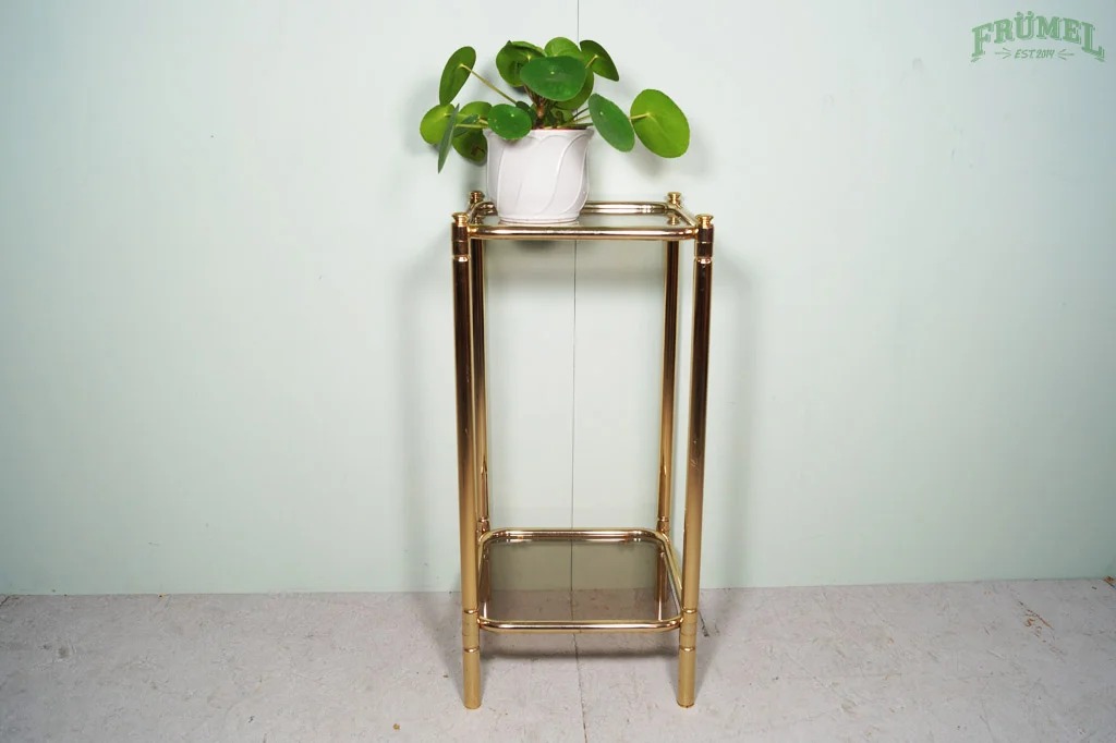 Gouden plantenstandaard met een witte pot en groene planten, op een lichte muur met groene tekst in de hoek.