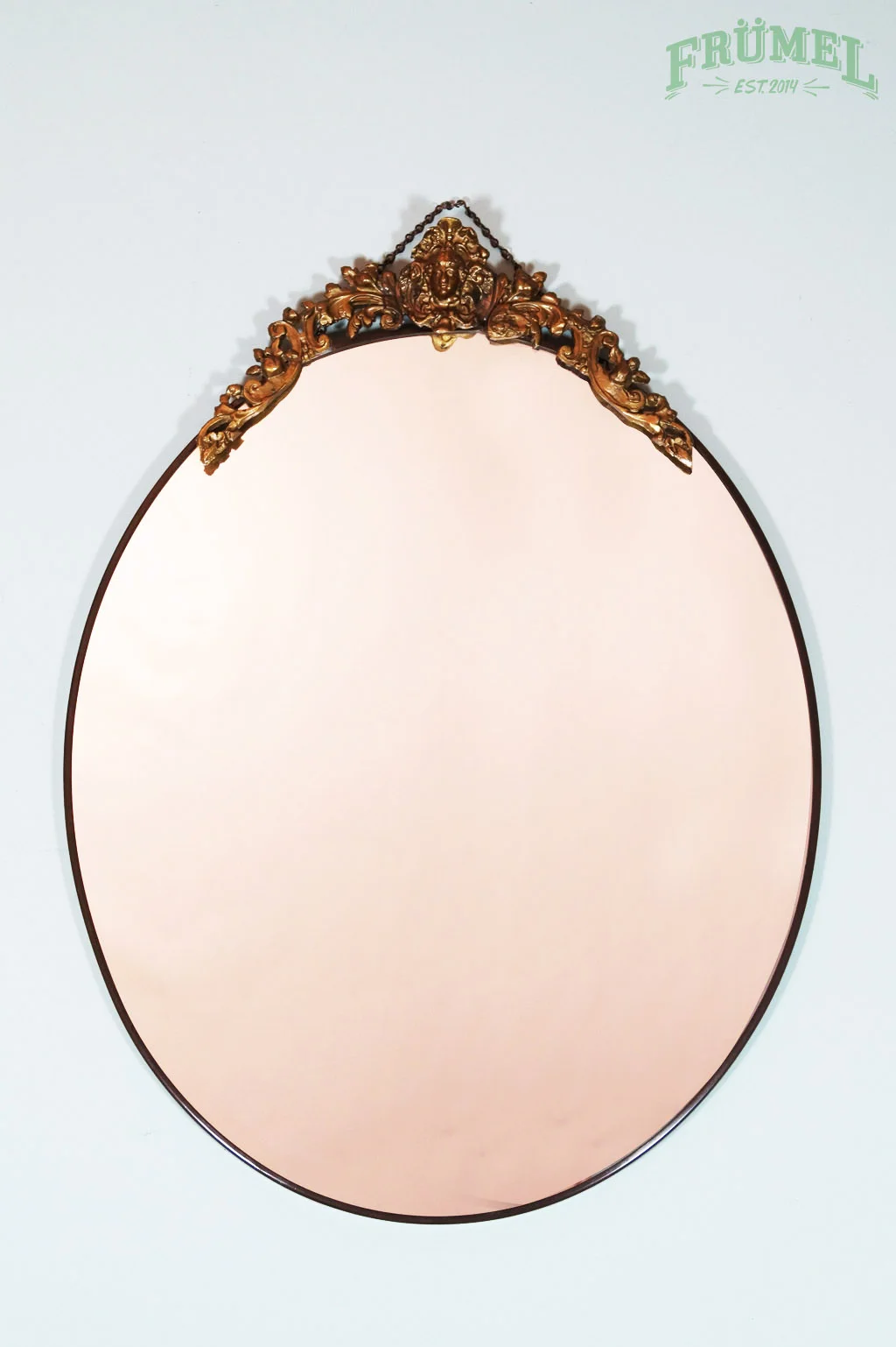 A decorative oval mirror with an ornate gold frame hanging on a light-colored wall, with a green logo in the top right corner that says "FRÜMEL EST. 2014."