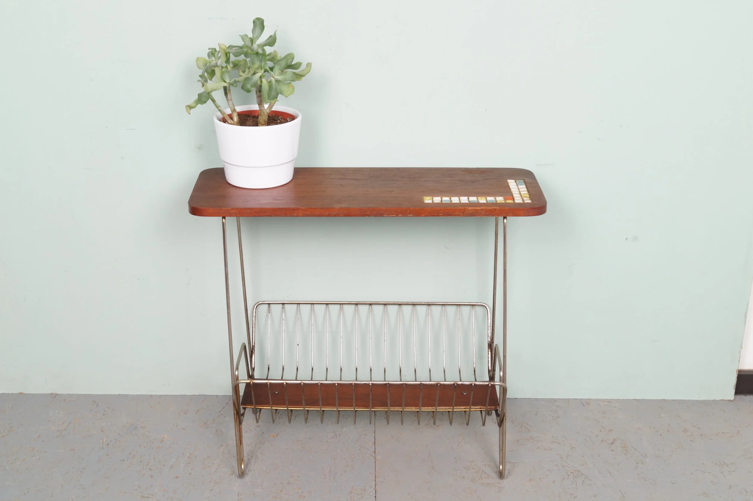 Houten tafel met metalen onderstel en een plantenpot met een plant erop, tegen een lichtgroene muur.