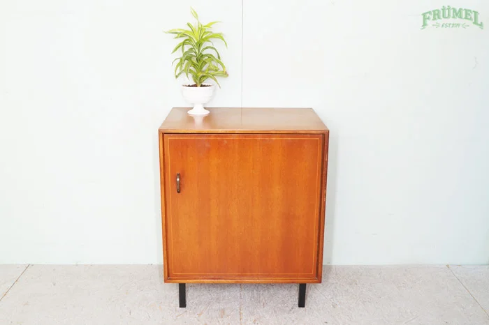 Een houten kast met een groene plant op een witte sierpot op de bovenkant, tegen een lichte muur met groene tekst.