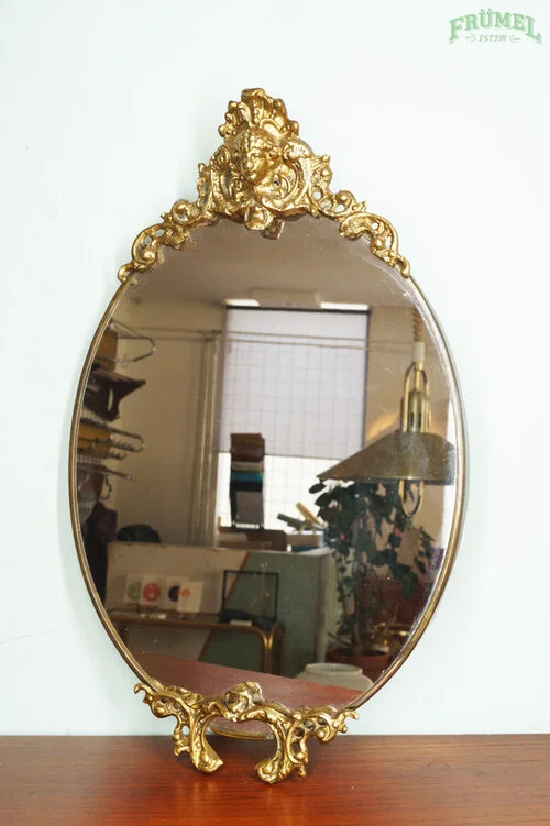 Ovaal spiegel met een gouden, versierde rand en koperen decoraties boven en onder, bevestigd aan een witte muur.