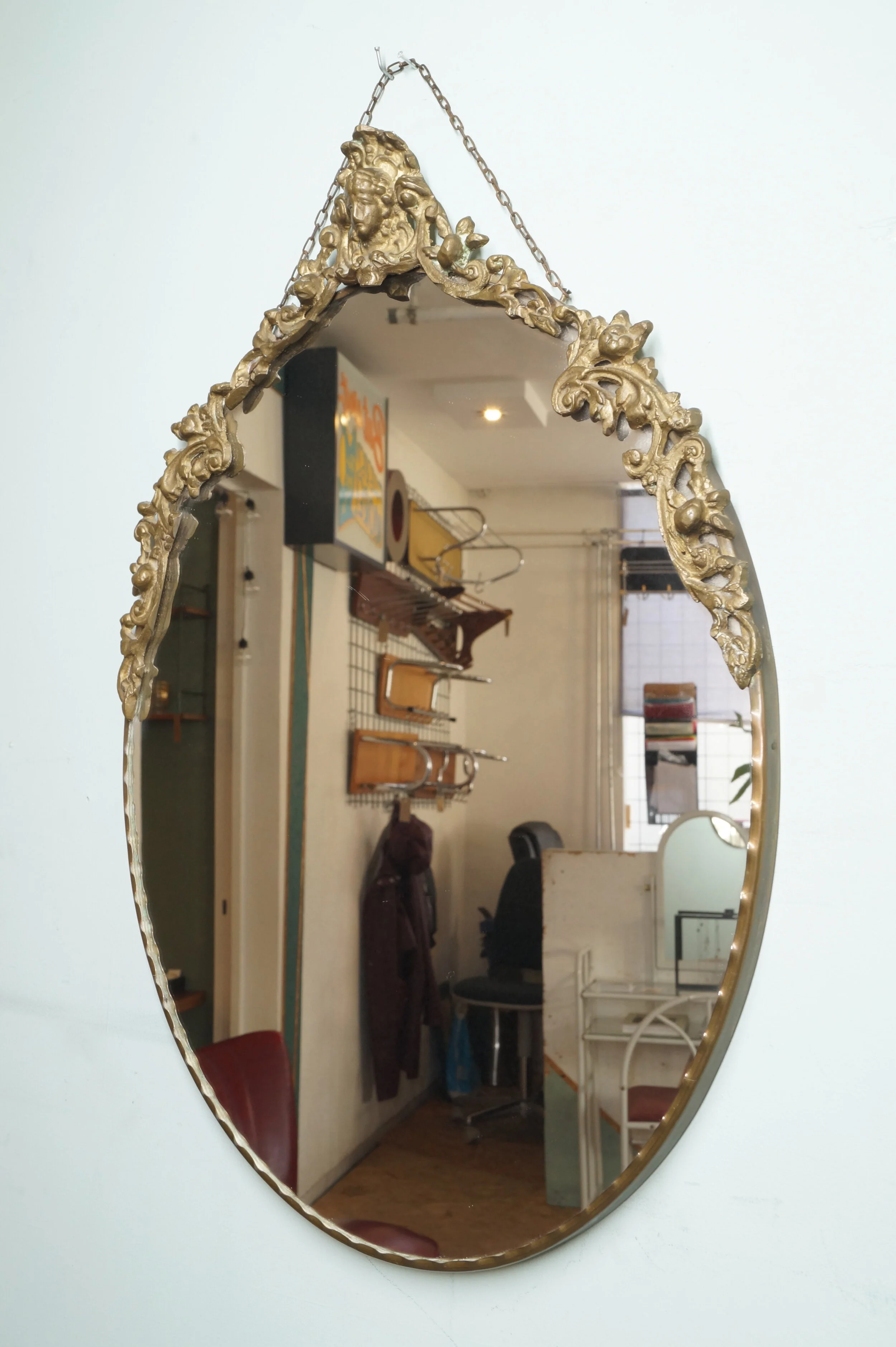 Decoratief gouden spiegel met ornamenteel frame hangt aan een witte muur, reflecteert een interieur met kantoormeubelen en een raam.