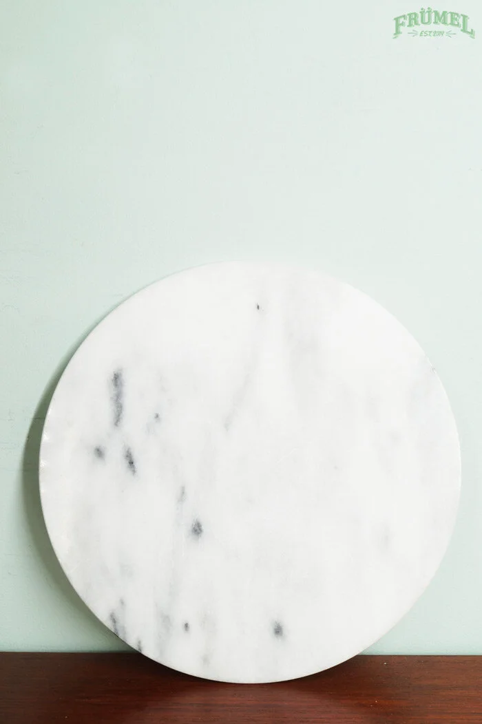 Ronde witte marmeren plaat met zwarte aders, tegen een lichtgroene muur, met een logo 'FRÜMEL' in de rechterbovenhoek.