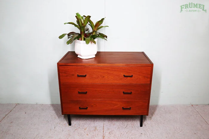 Een houten ladekast met drie lades en zwarte handgrepen, met een groene plant in een witte pot erop, tegen een witte muur met een groene logo in de hoek.