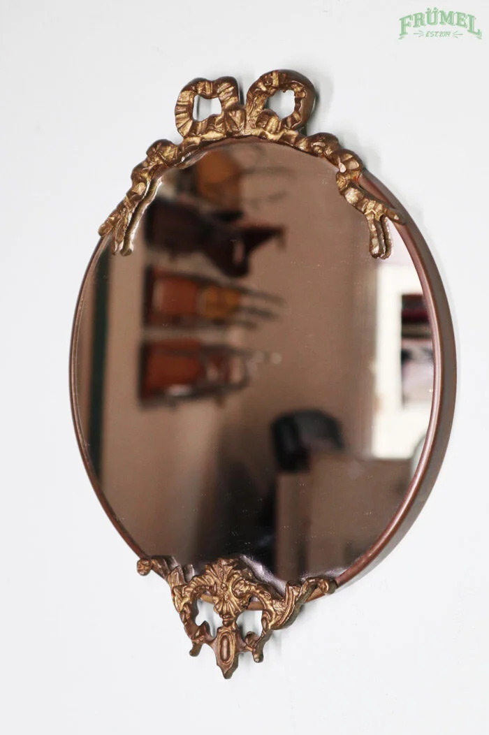 Een ovalen spiegel met een ornamentele gouden lijst hangt aan een witte muur, reflecteert een kamer met boekenplanken en een bureau.
