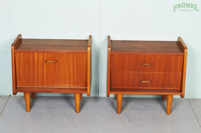 Twee houten nachtkastjes met drie poten en metalen handgrepen, naast elkaar tegen een witte muur.
