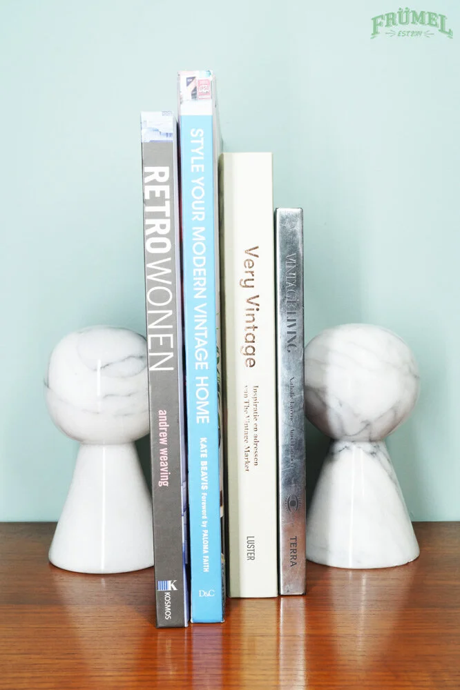 Vijf boeken staan rechtop op een houten tafel tussen twee witte marmeren sculpturen, tegen een lichtgroene muur.