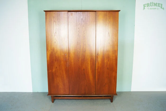 Een oude houten Louis van Teeffelen kledingkast met drie deuren, staand tegen een lichtgroene muur en een witte muur. Rechts staat een deel van een groene tekst die zegt "FRÜMEL".