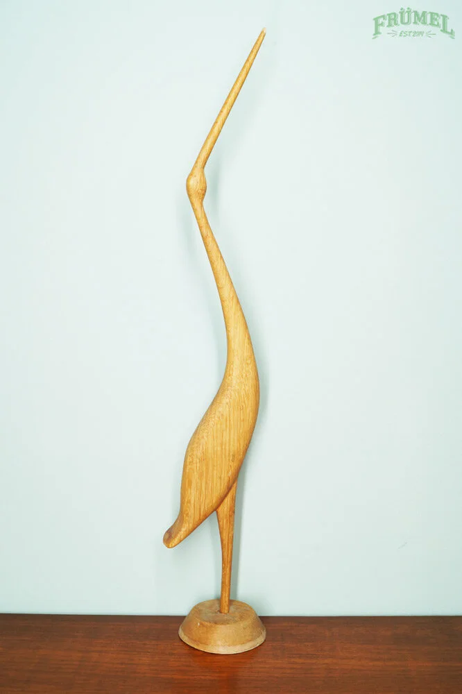 Houten decoratie in de vorm van een vogel met een lange, kromme nek, geplaatst op een houten ondergrond. Logo 'FRÜMEL' zichtbaar in de bovenhoek rechts.
