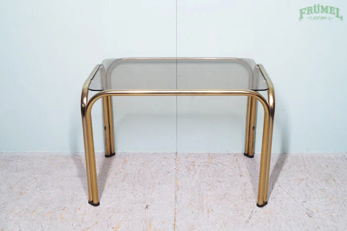 Een tafel met een glazen blad en gouden metalen poten, geplaatst tegen een witte muur met een logo dat 'FRÜMEI' zegt.