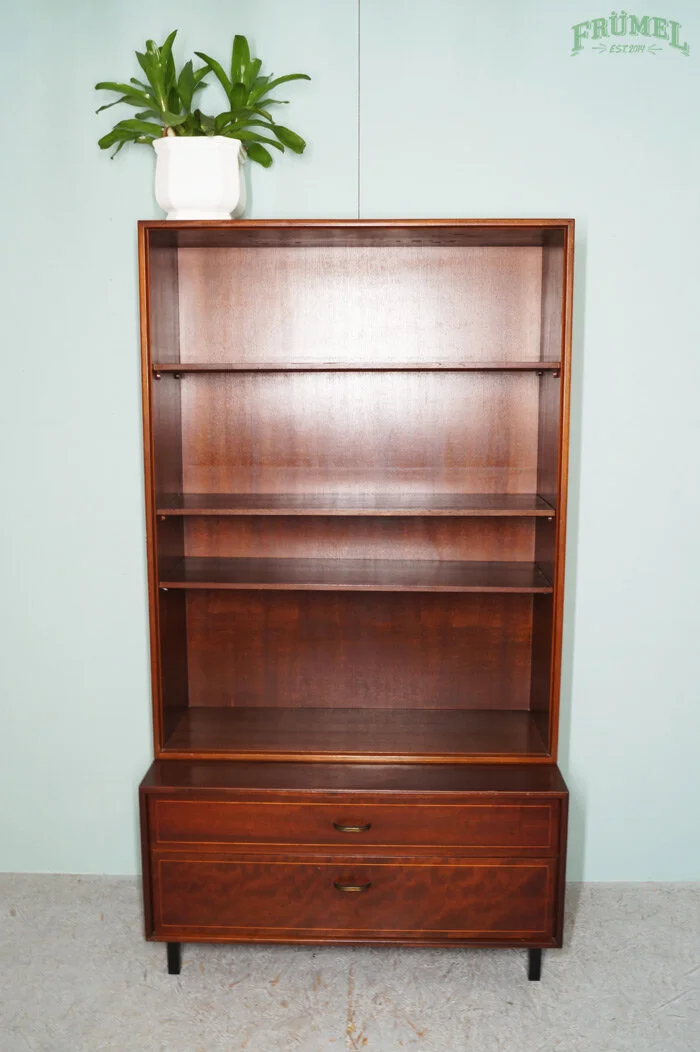 Een lege houten boekenkast met drie planken en twee lades onderaan, met een witte pot met groene plant op de bovenkant, tegen een lichtblauwe muur.