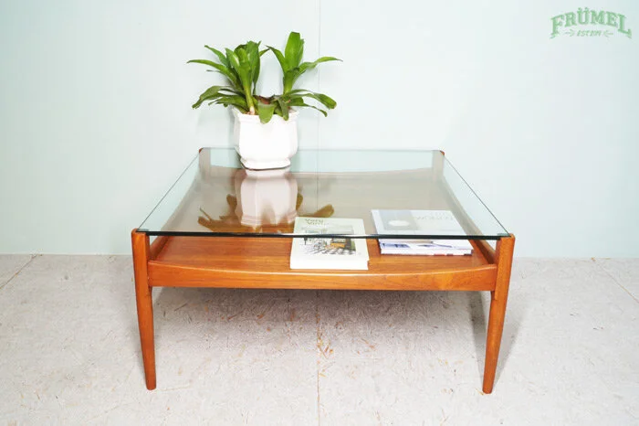 Houten salontafel met glazen blad en een witte plantenpot met groene bladeren, enkele tijdschriften en een brochure erop, tegen een lichtgroene muur met groene tekst.