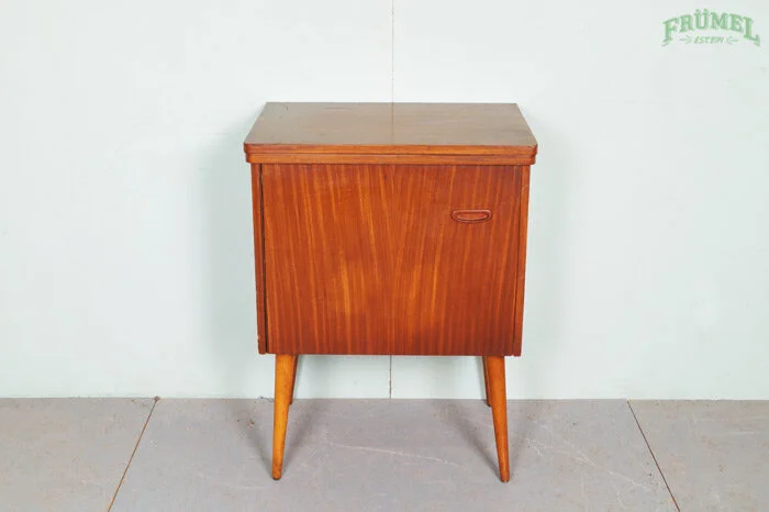 Een vintage houten kast met schuifdeur en afneembare poten, geplaatst tegen een witte muur.
