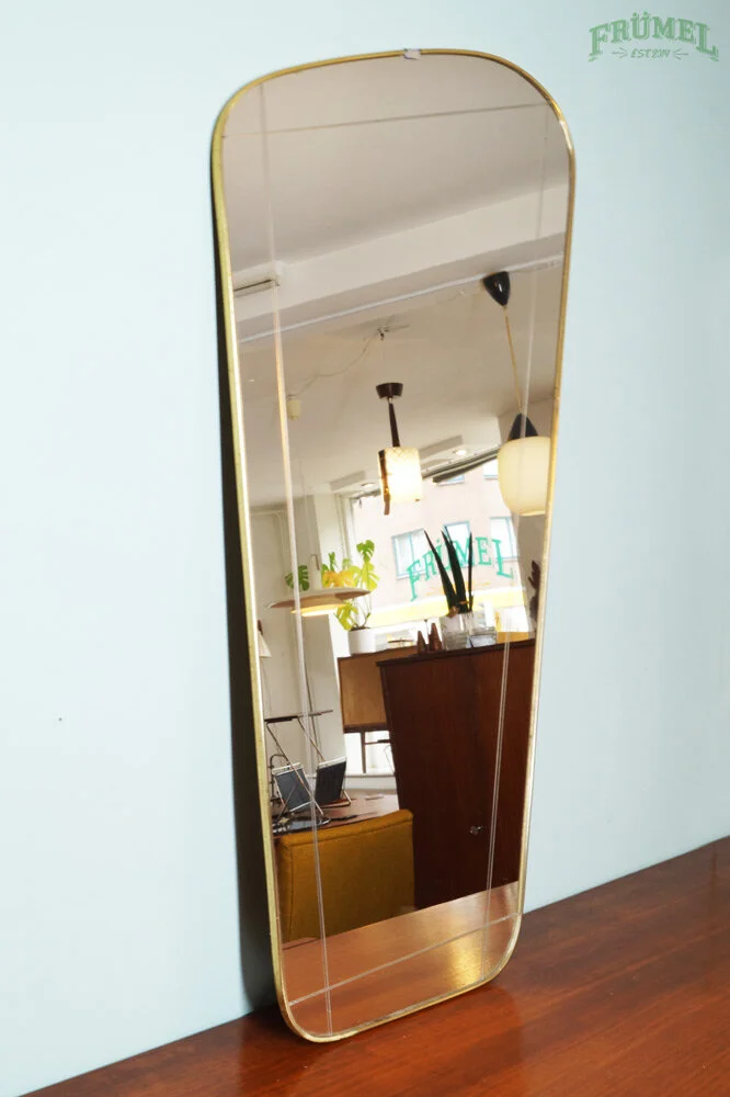 Een verticale spiegel met een gouden rand staat tegen een lichte blauwe muur, die de binnenkant van een moderne, lichte kamer reflecteert.