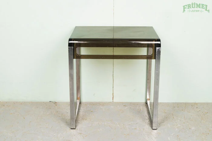 Een metalen tafel met een zwart bovenblad, staand tegen een lichte muur met een groen logo in de rechterbovenhoek.