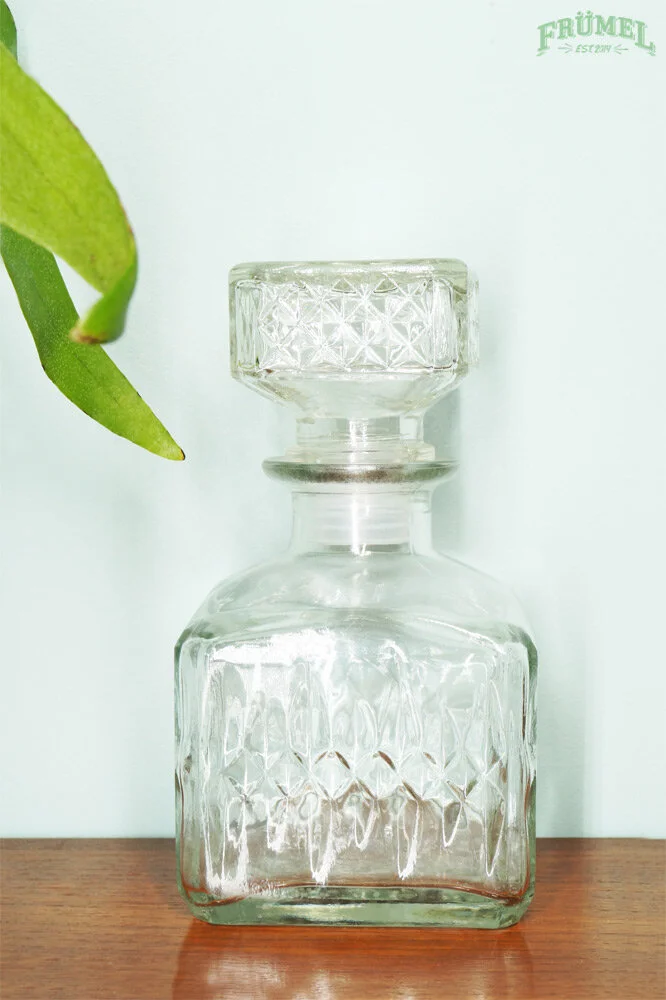 Kristal heldere drinkfles met geometrisch patroon op houten oppervlak, groene bladeren in de linkerhoek, lichte achtergrond, boven rechts tekst 'FRÜMEL EST. 2017'