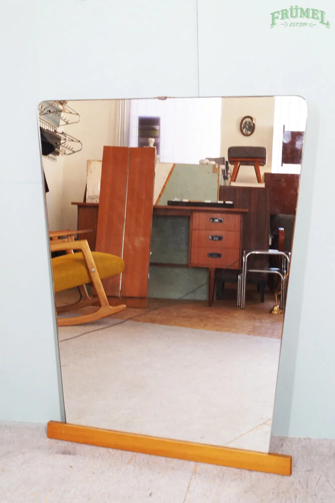 Een staande spiegel met een houten frame, weerspiegelt een interieur met vintage meubels zoals een groene en houten kast, een gele hangstoel en andere houten meubels.