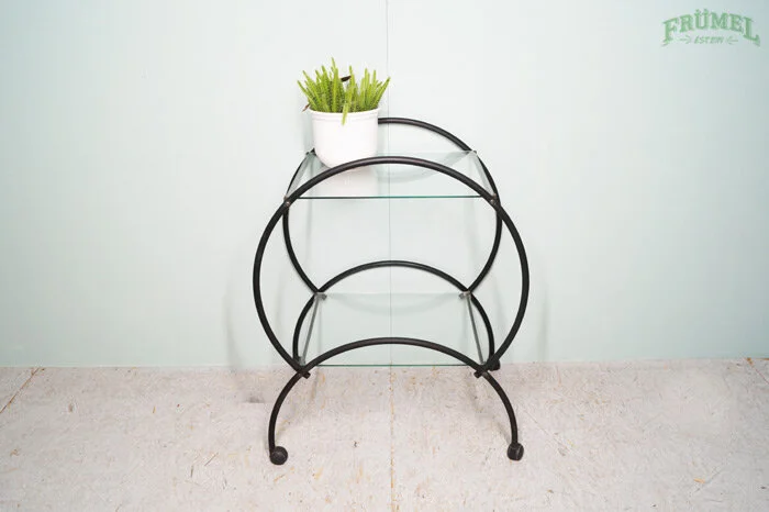 Een metalen plantenstandaard met drie ronde bakken, met een plantenpot met groen blad op de bovenste plank, tegen een witte muur met een groene tekst 'FRÜMET'.