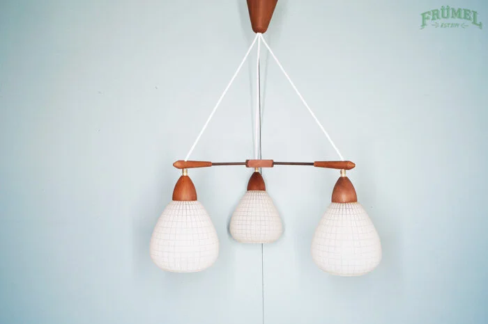 Een hanglamp met drie witte kappen met een rasterpatroon, bevestigd aan een houten steun en opgehangen aan witte touwen tegen een lichtblauwe muur.