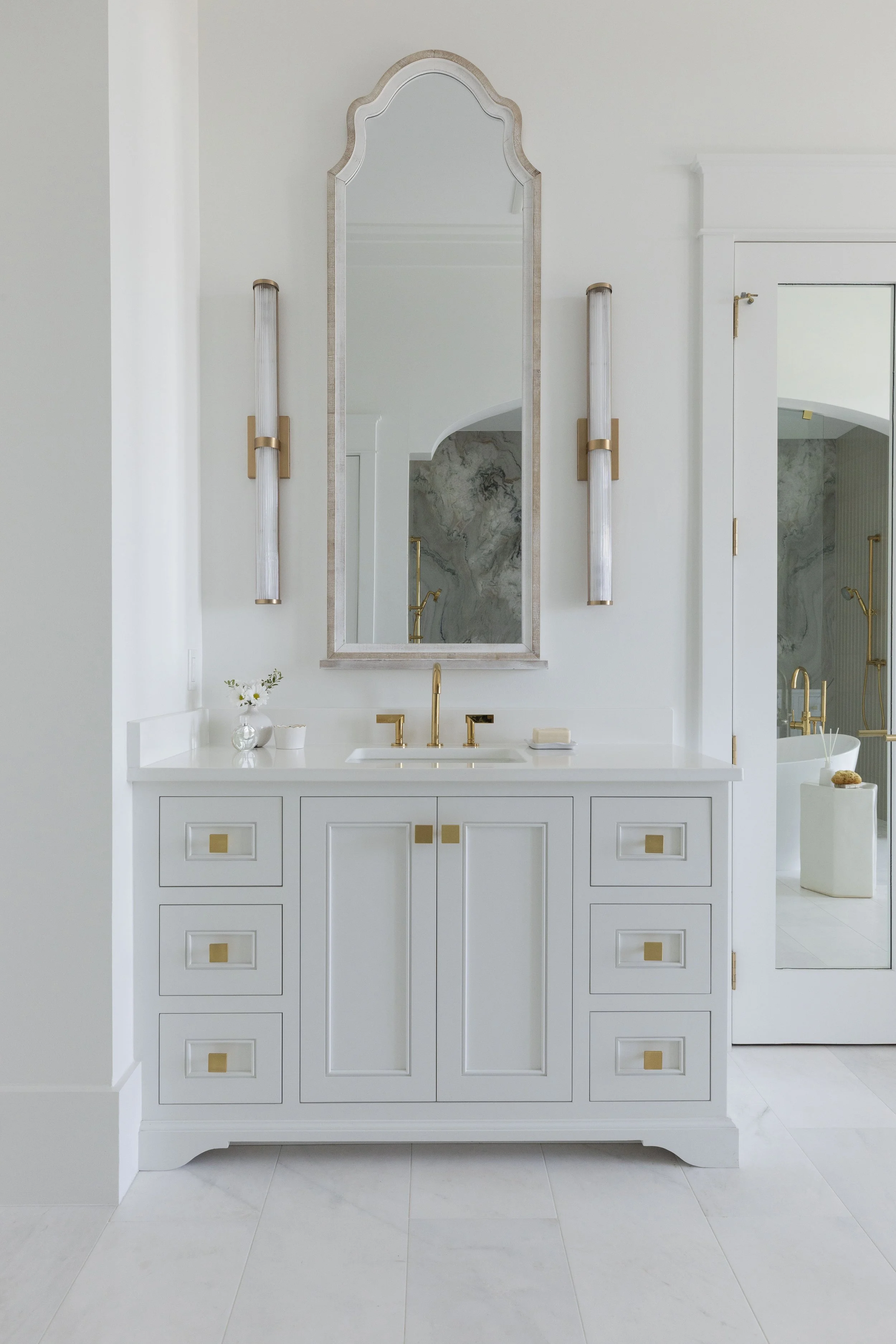 Custom white bathroom vanity in Blue Mountain Beach by Cindy McCarley Designs, featuring brass hardware, an arched mirror and fluted brass sconces.