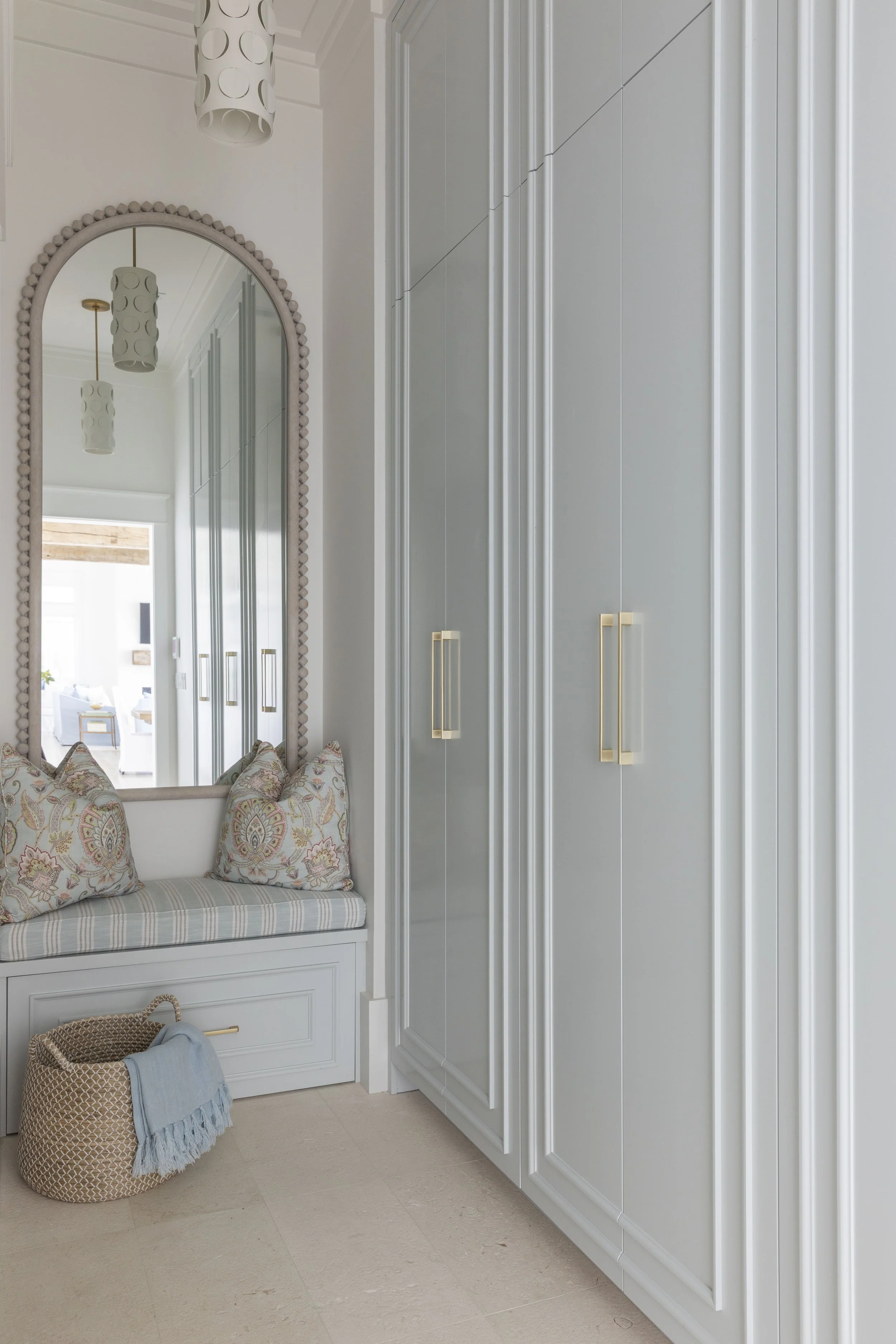 Custom built-in storage in a Blue Mountain Beach home by Cindy McCarley Designs, featuring soft blue cabinetry, brass hardware, a cushioned bench and arched mirror.