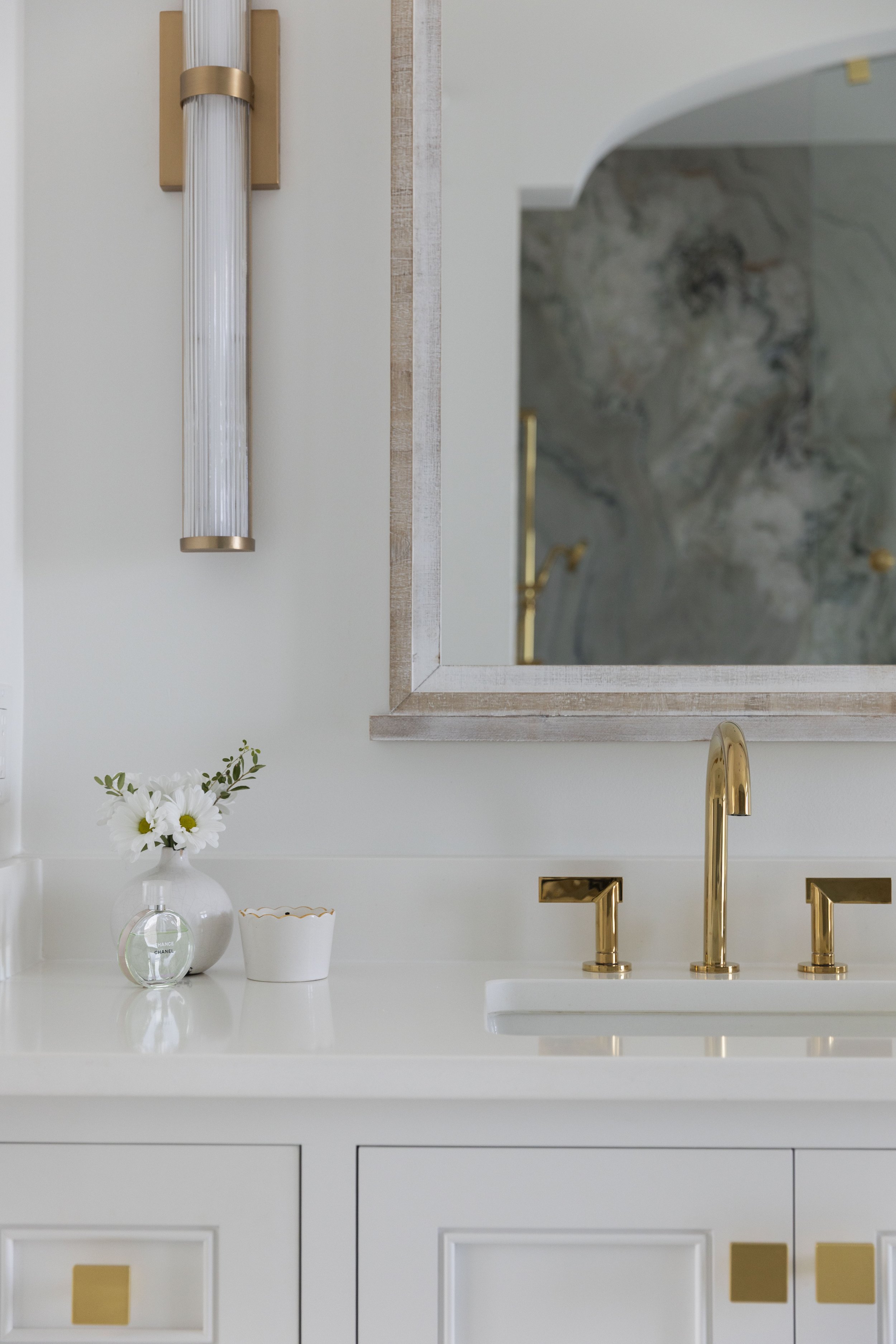 Close-up of a custom bathroom vanity by Cindy McCarley Designs featuring polished brass fixtures, white cabinetry, an elegant framed mirror and brass wall sconce.