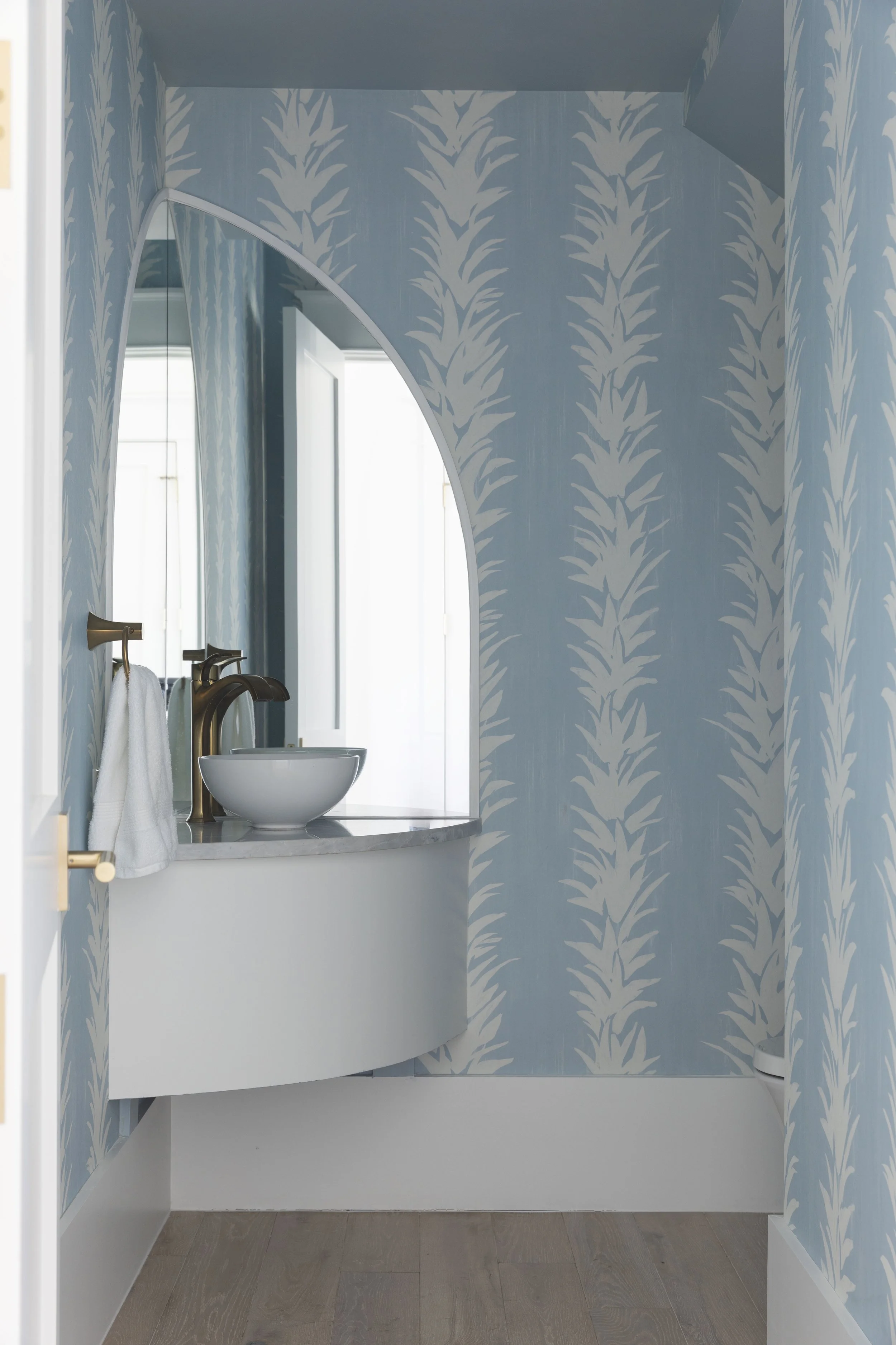Blue and white powder room in a Blue Mountain Beach home by Cindy McCarley Designs, featuring botanical wallpaper, an arched mirror and brass fixtures