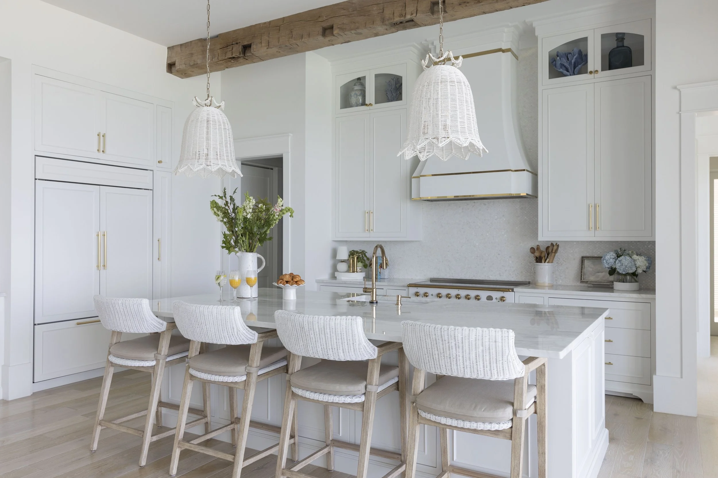 Bright coastal kitchen in Blue Mountain Beach by Cindy McCarley Designs, featuring white cabinetry, brass accents, woven pendant lights and a large marble island.