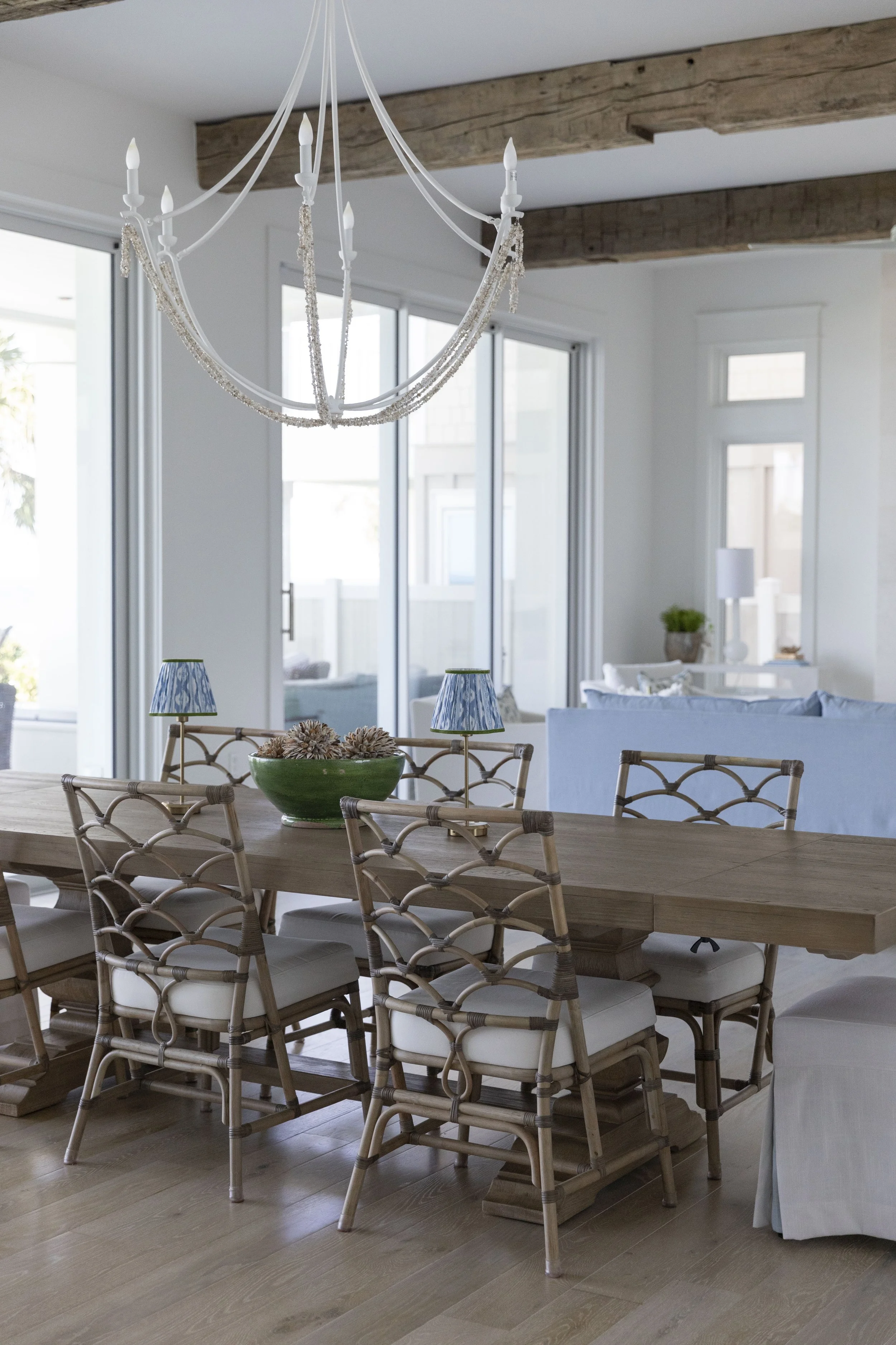 Coastal dining room in Blue Mountain Beach by Cindy McCarley Designs, featuring a natural wood table, woven dining chairs, statement chandelier and reclaimed wood beams.