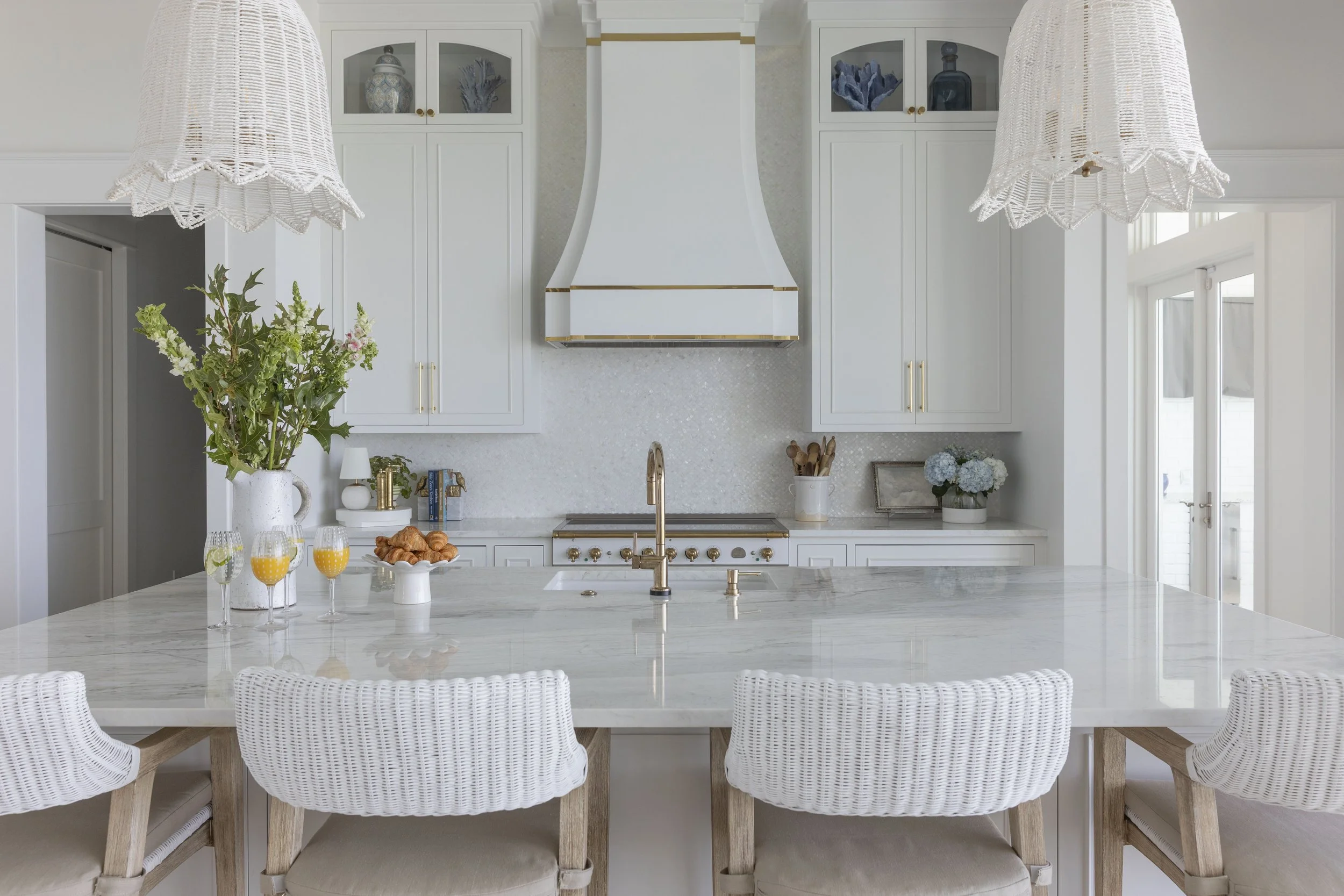 White coastal kitchen in Blue Mountain Beach by Cindy McCarley Designs, featuring an oversized marble island, woven barstools, brass fixtures and statement pendant lights.
