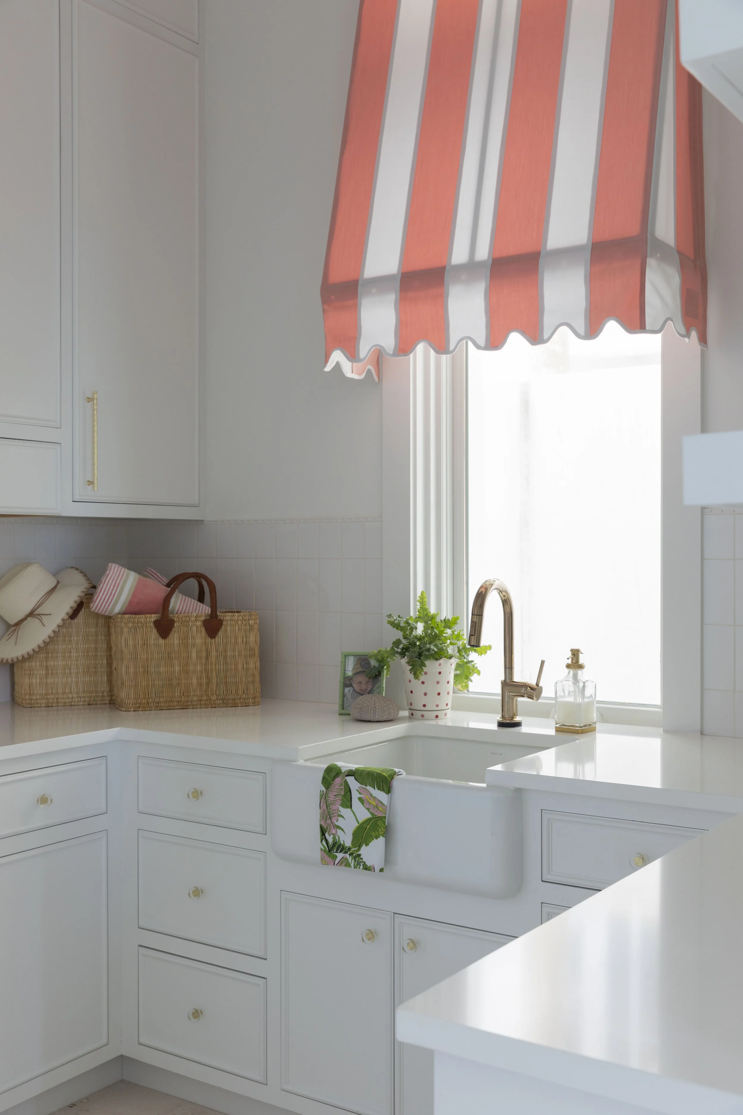 Bright coastal laundry room in Blue Mountain Beach by Cindy McCarley Designs, featuring white cabinetry, farmhouse sink, brass fixtures and a coral-and-white striped window shade.