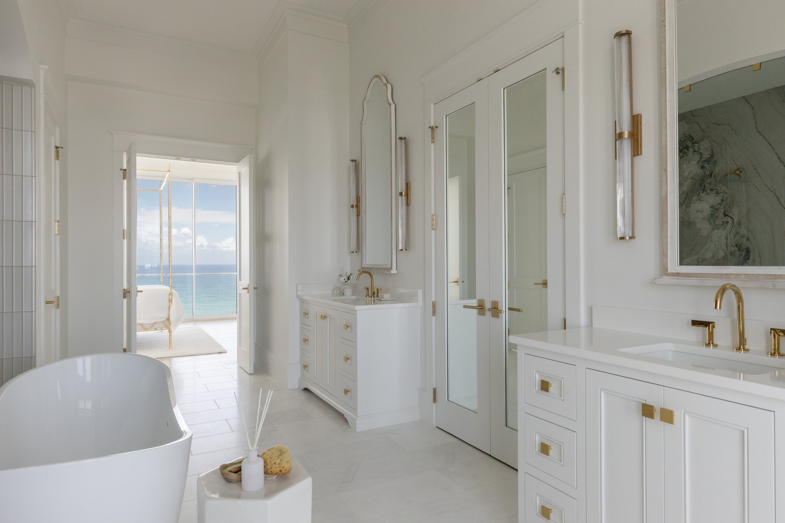 Spacious primary bathroom in Blue Mountain Beach by Cindy McCarley Designs, featuring custom white vanities, brass accents, a freestanding tub and views into the Gulf-front bedroom.