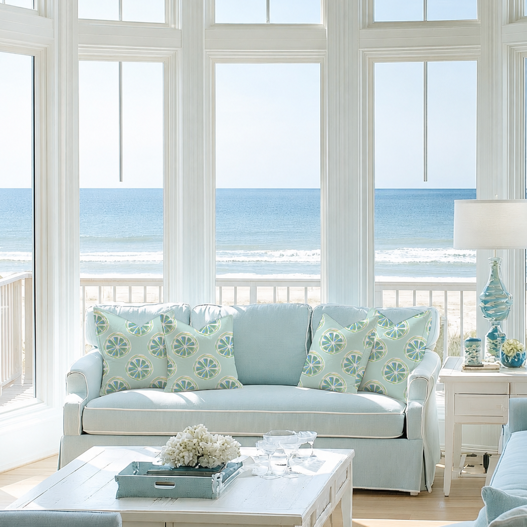 Living room with light blue sofa, patterned cushions, white wooden coffee table with flowers and glasses, ocean view through large windows, beach and sky outside.