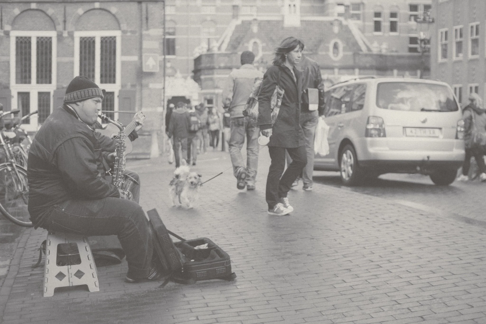 A street scene with a man playing the saxophone on a small stool, a woman walking her dog, and several pedestrians. Cars are parked along the street and buildings line the background.