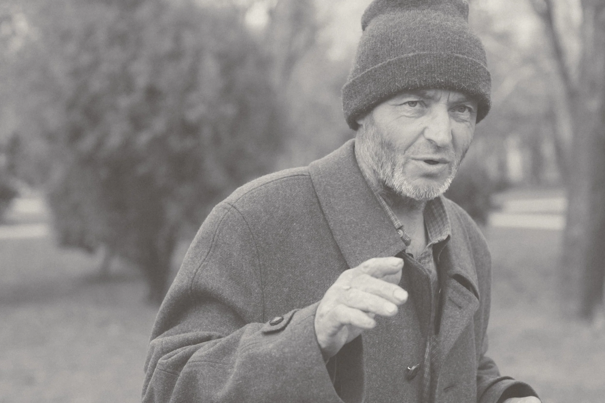A black-and-white photo of a man wearing a knit cap and coat, standing outdoors, with trees and a park in the background.