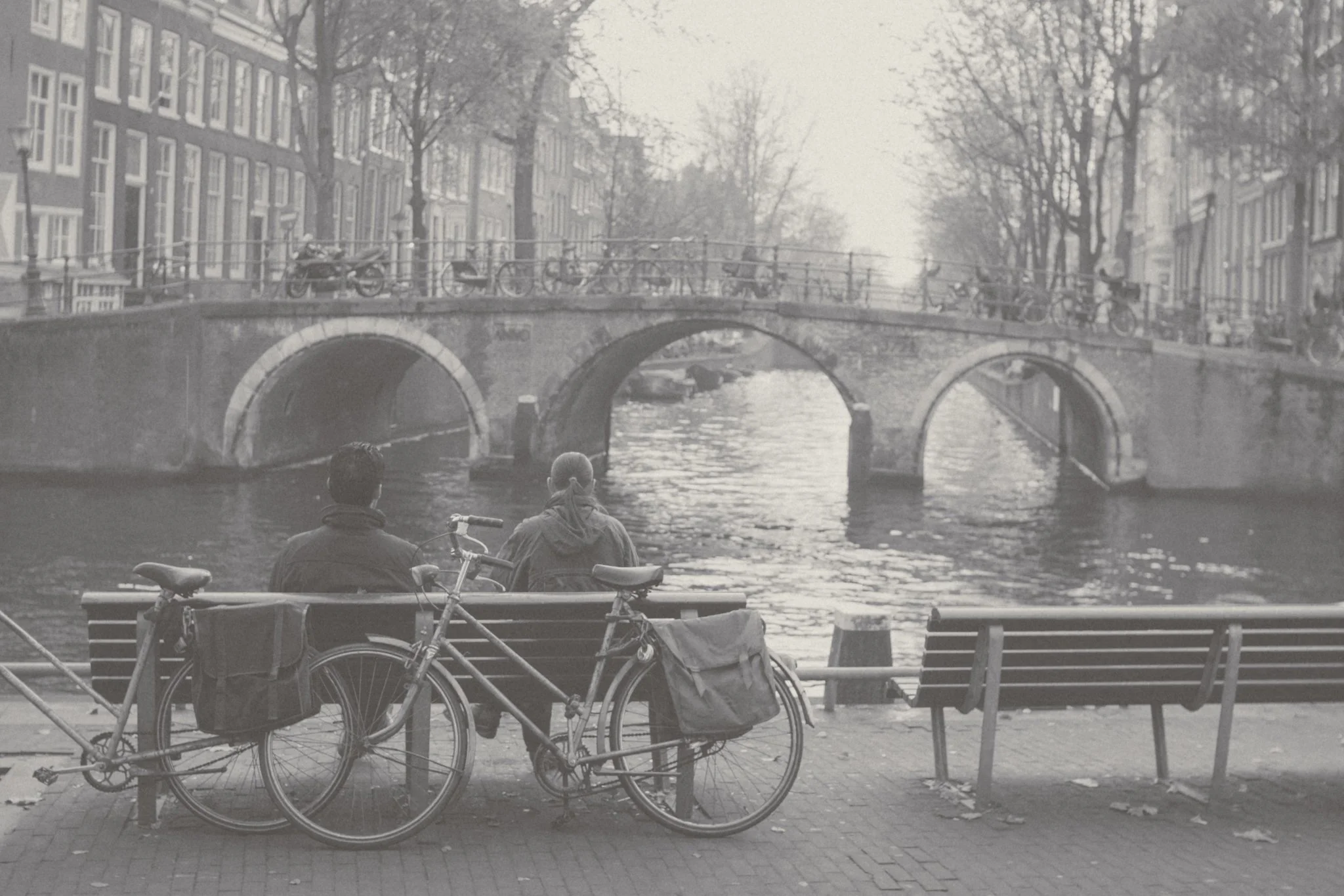 Two men are sitting on a bench by a canal in a city, with a bicycle parked next to them. A bridge and old buildings are visible in the background.