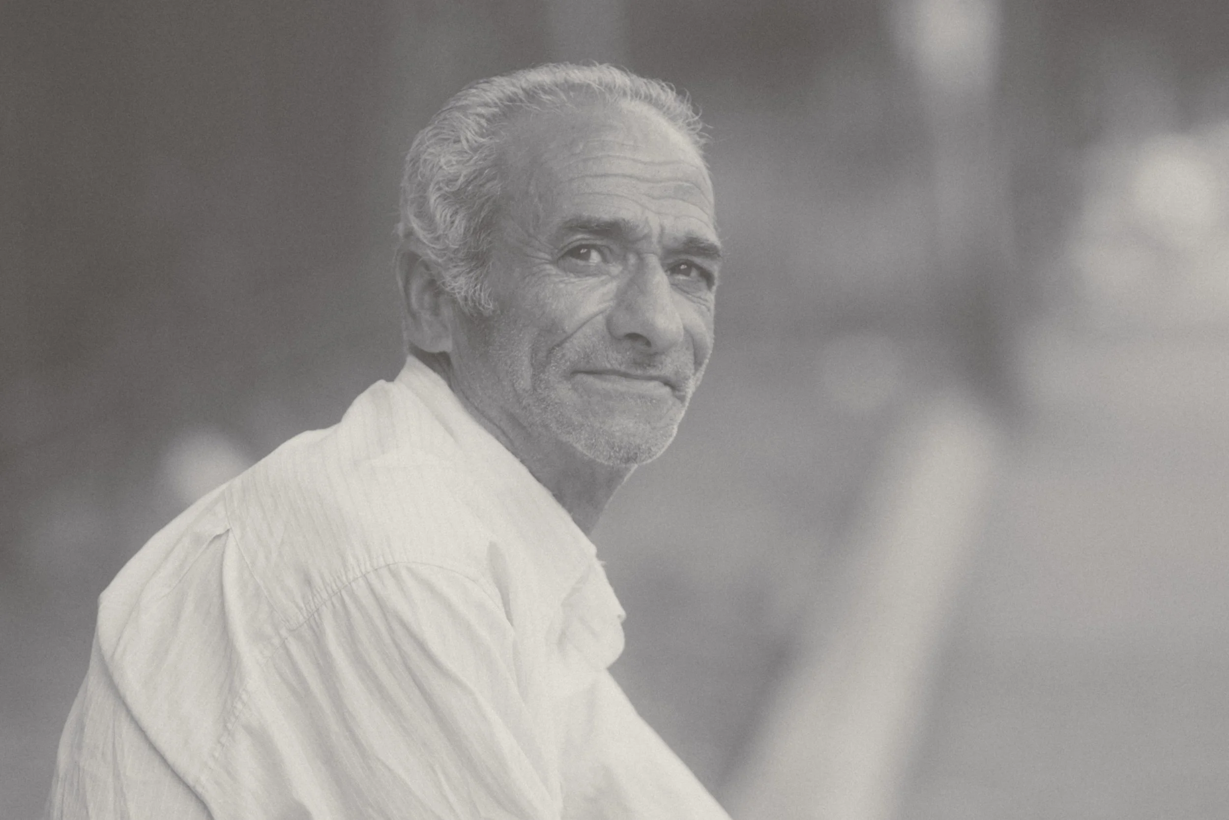 A black and white photo of an elderly man with gray hair, wearing a light-colored shirt, looking over his shoulder with a subtle smile.