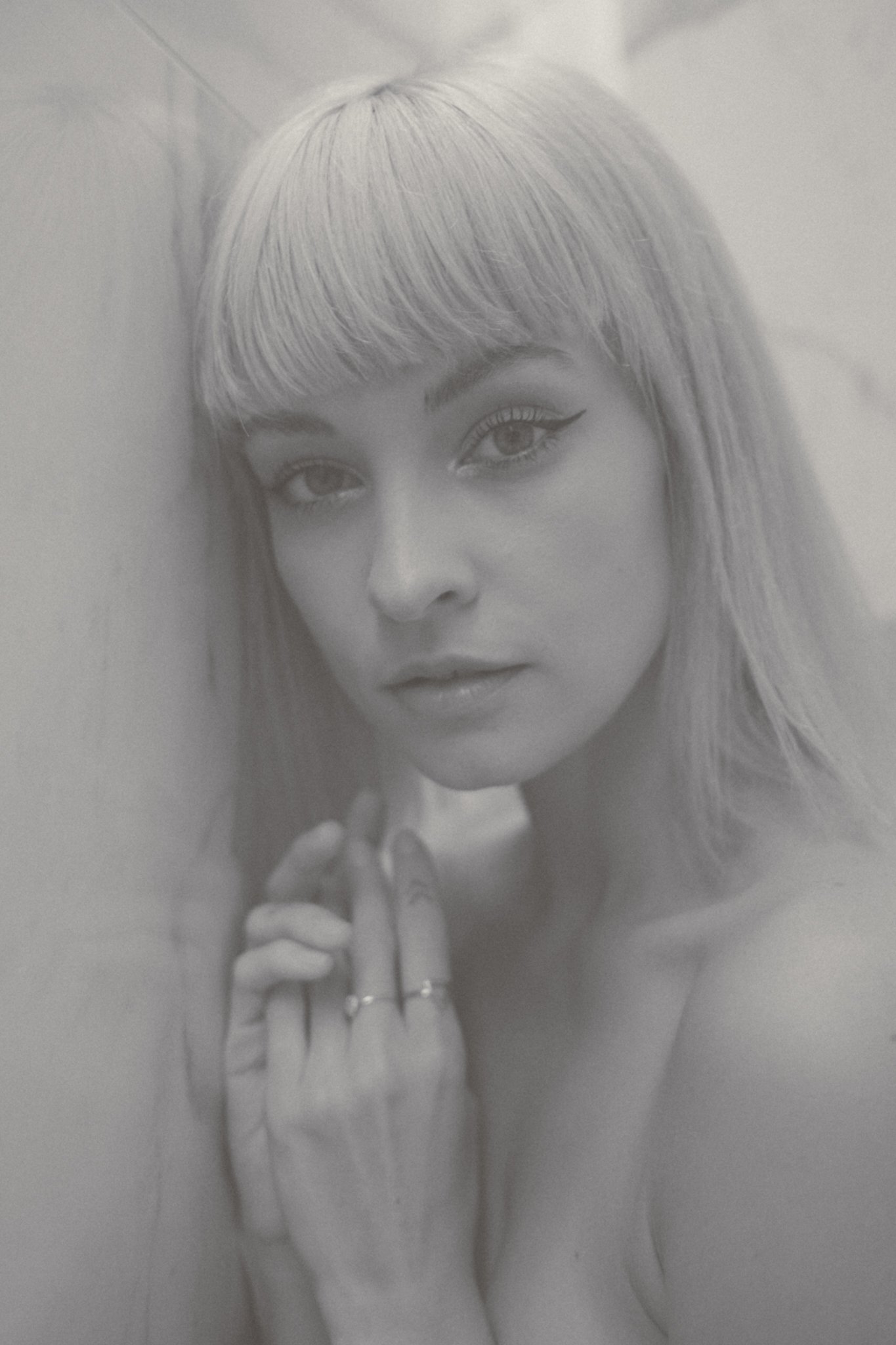 Black and white photo of a woman with long hair and bangs, looking directly at the camera, with makeup and a hand holding a mascara wand near her eye.